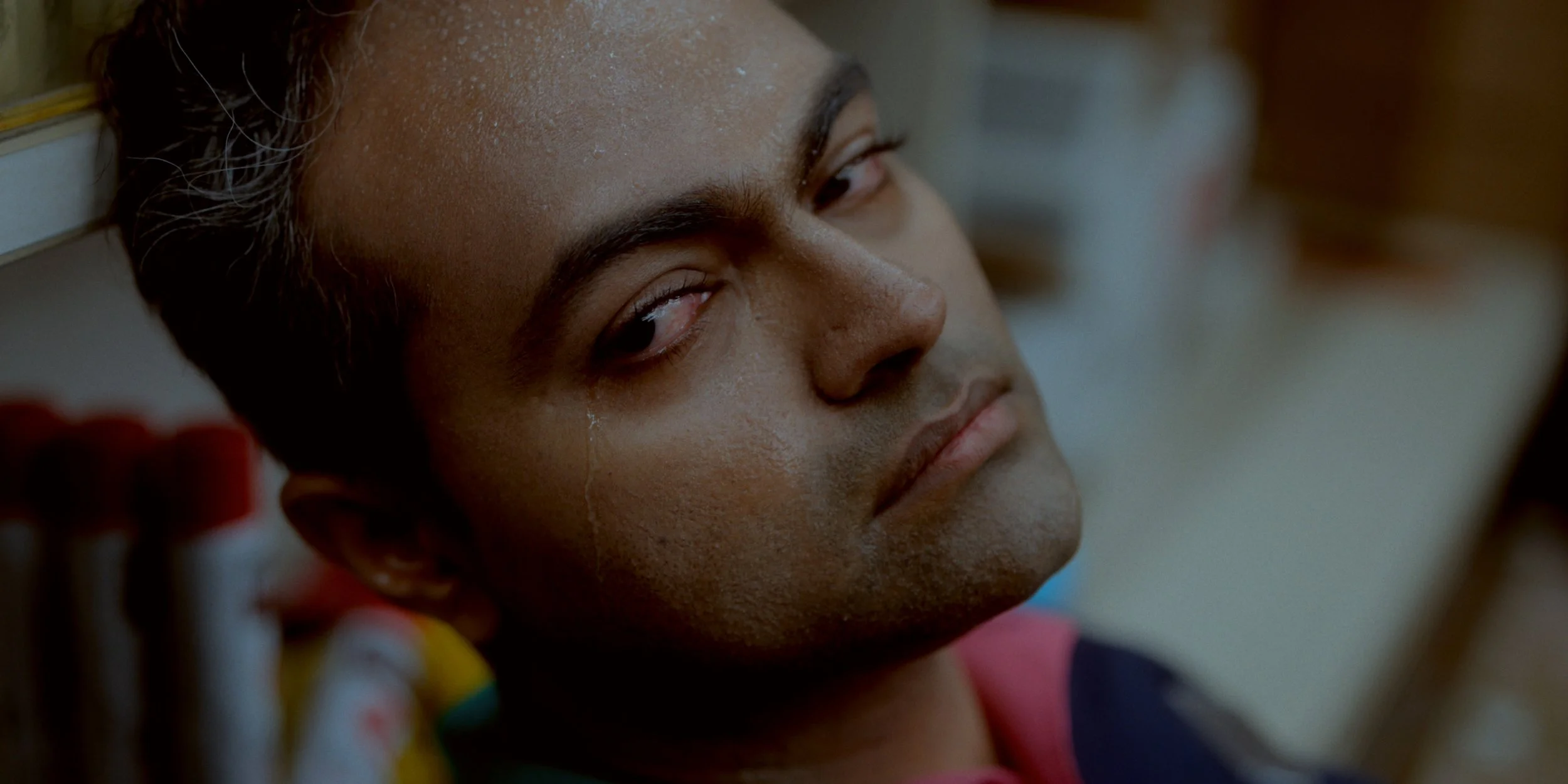 Close-up of a young man with a serious expression, lying down with a crack on his cheek, with objects in the background.