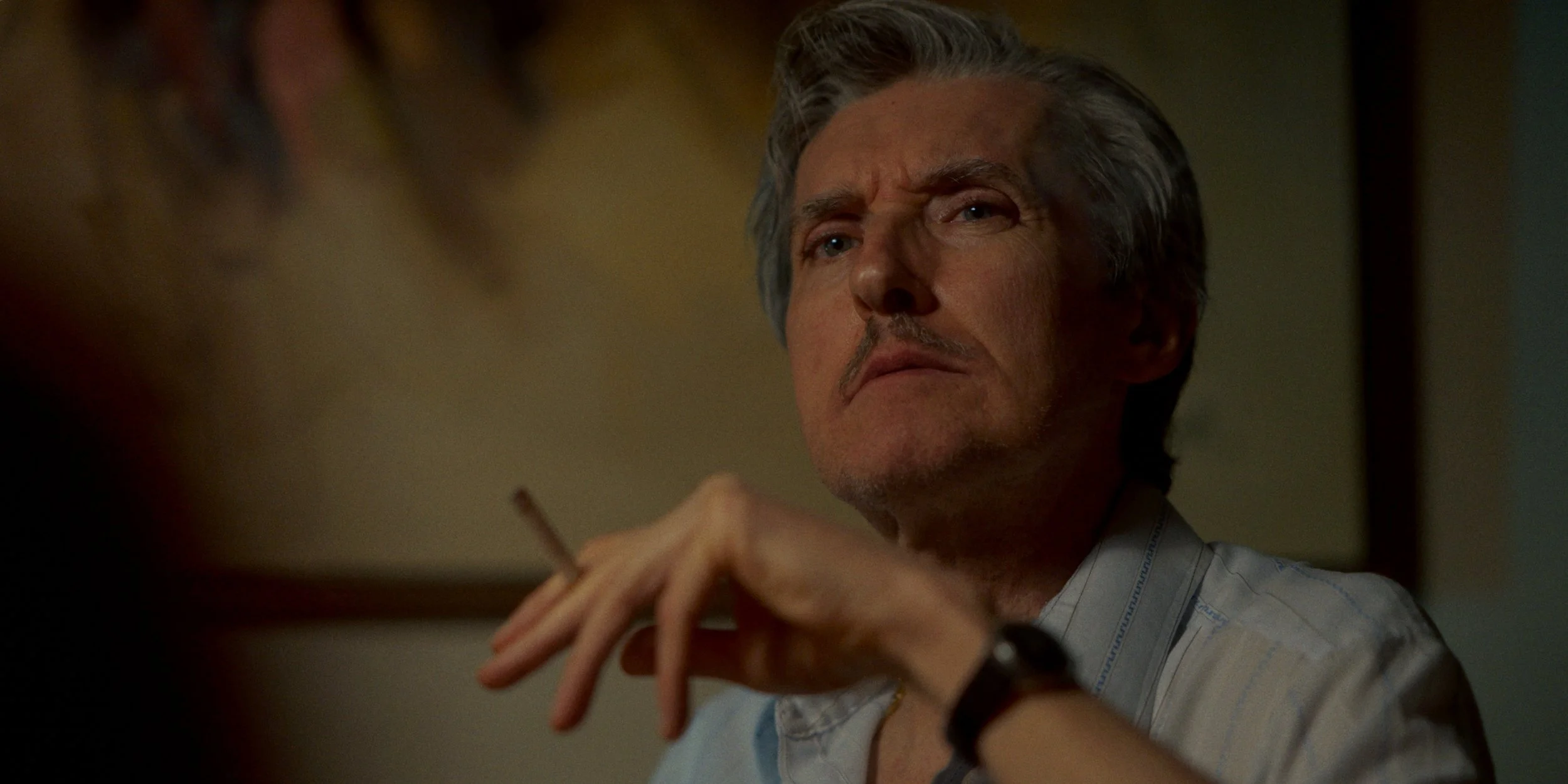 A middle-aged man with gray hair and a mustache gazes thoughtfully. He is wearing a light-colored shirt and has a watch on his wrist. His hand is near his face, holding a lit cigarette. The background is blurred, indicating an indoor setting.
