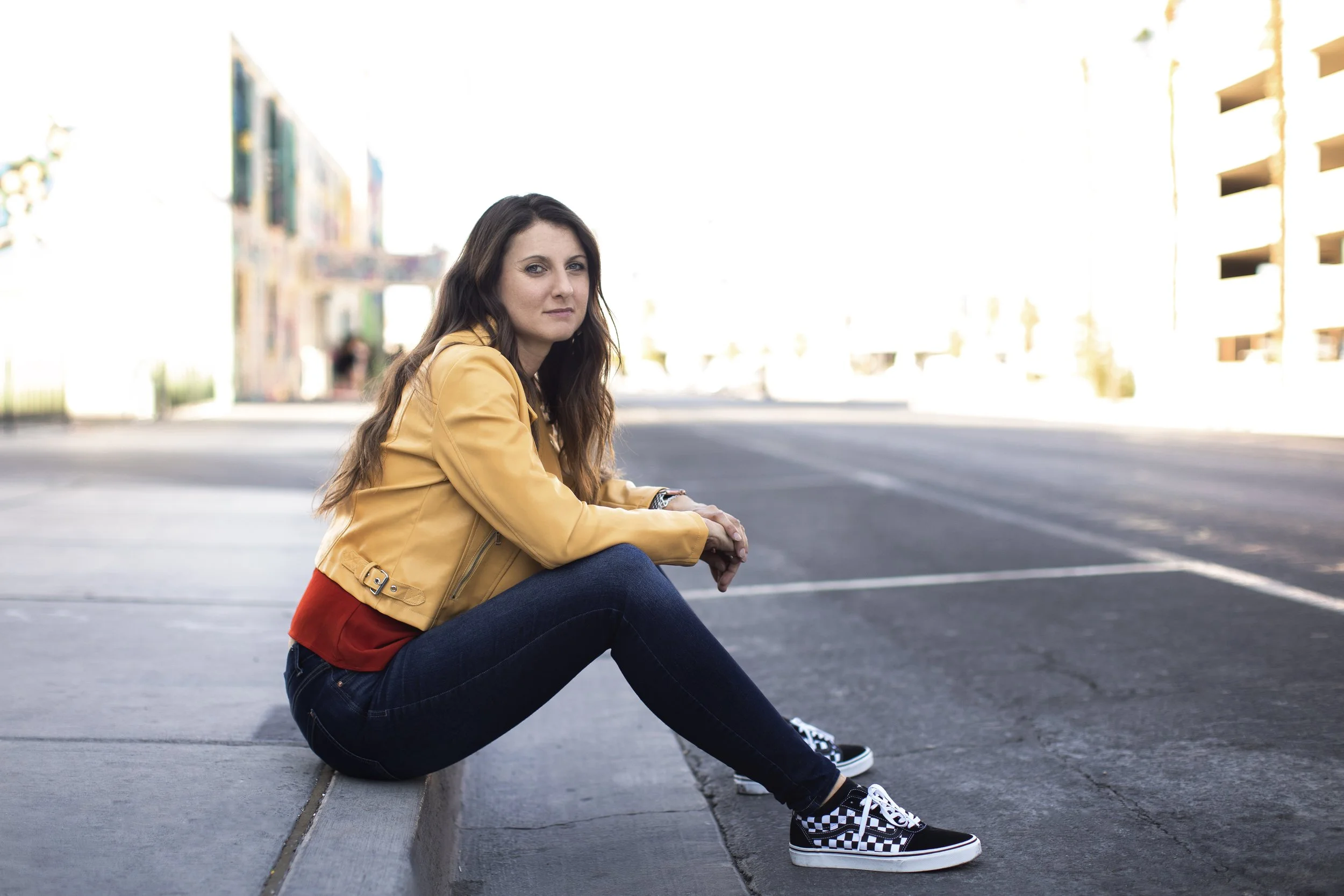 Young woman sitting on sidewalk in urban area, wearing yellow jacket, red shirt, black jeans, and checkerboard Vans sneakers, with buildings in the background.