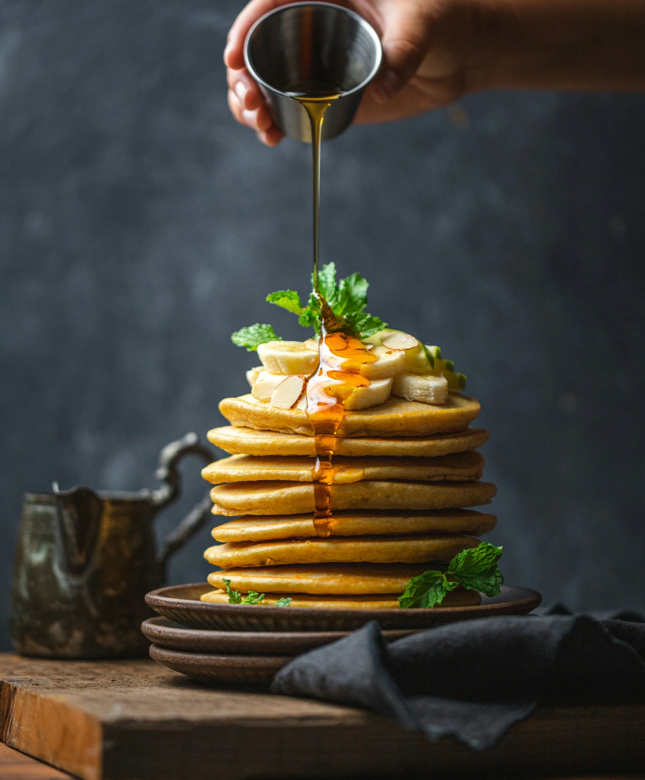 Stack of golden pancakes topped with sliced bananas, mint leaves, and syrup being poured from a small pitcher, set on a dark wooden table with a dark background.