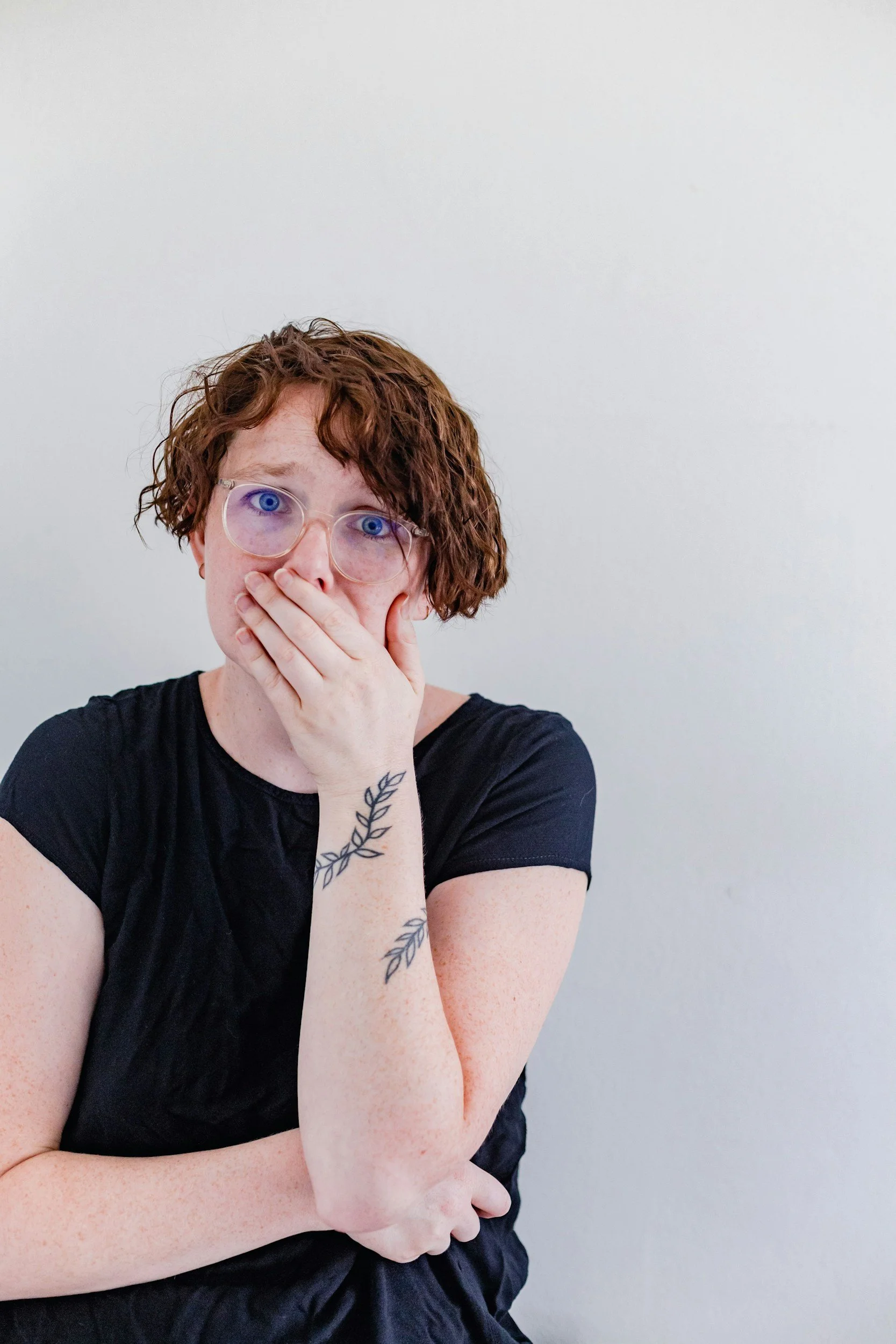 A woman with short curly reddish-brown hair and glasses covering her mouth with her hand, wearing a black shirt, standing against a light-colored wall.