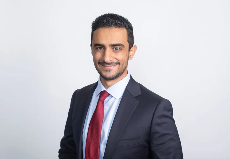Professional headshot of a man in a dark suit and red tie on a white background