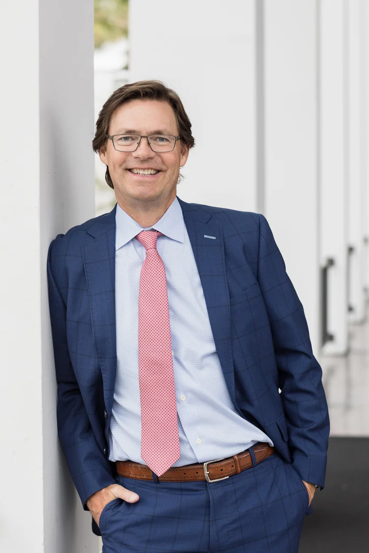 Modern headshot of a doctor in glasses and a blue suit leaning against a white column