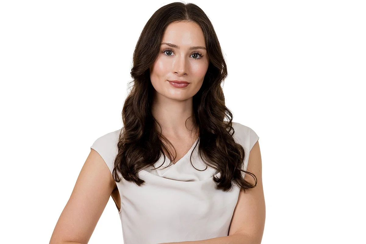 A woman with long, wavy brown hair, wearing a white sleeveless blouse, looking at the camera with a slight smile.