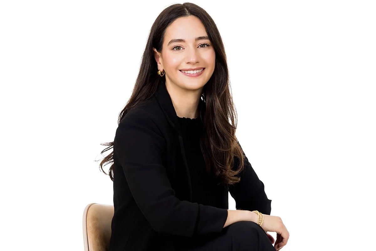 A young woman with long brown hair, wearing a black jacket and gold earrings, smiling and sitting on a beige chair against a white background.