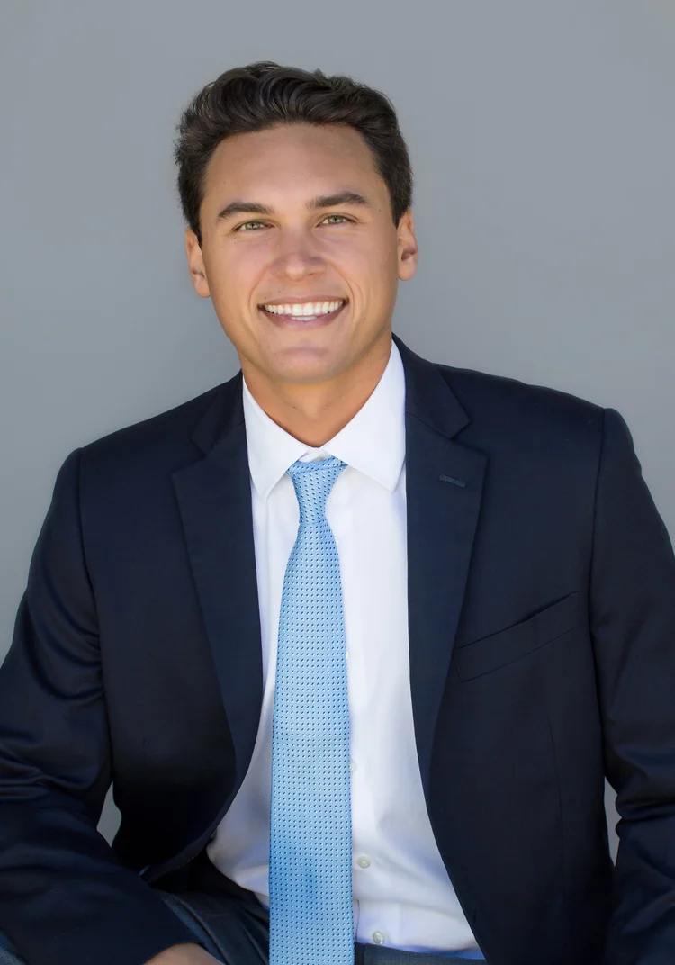 Studio headshot of a young professional in a navy suit and light blue tie on a grey background