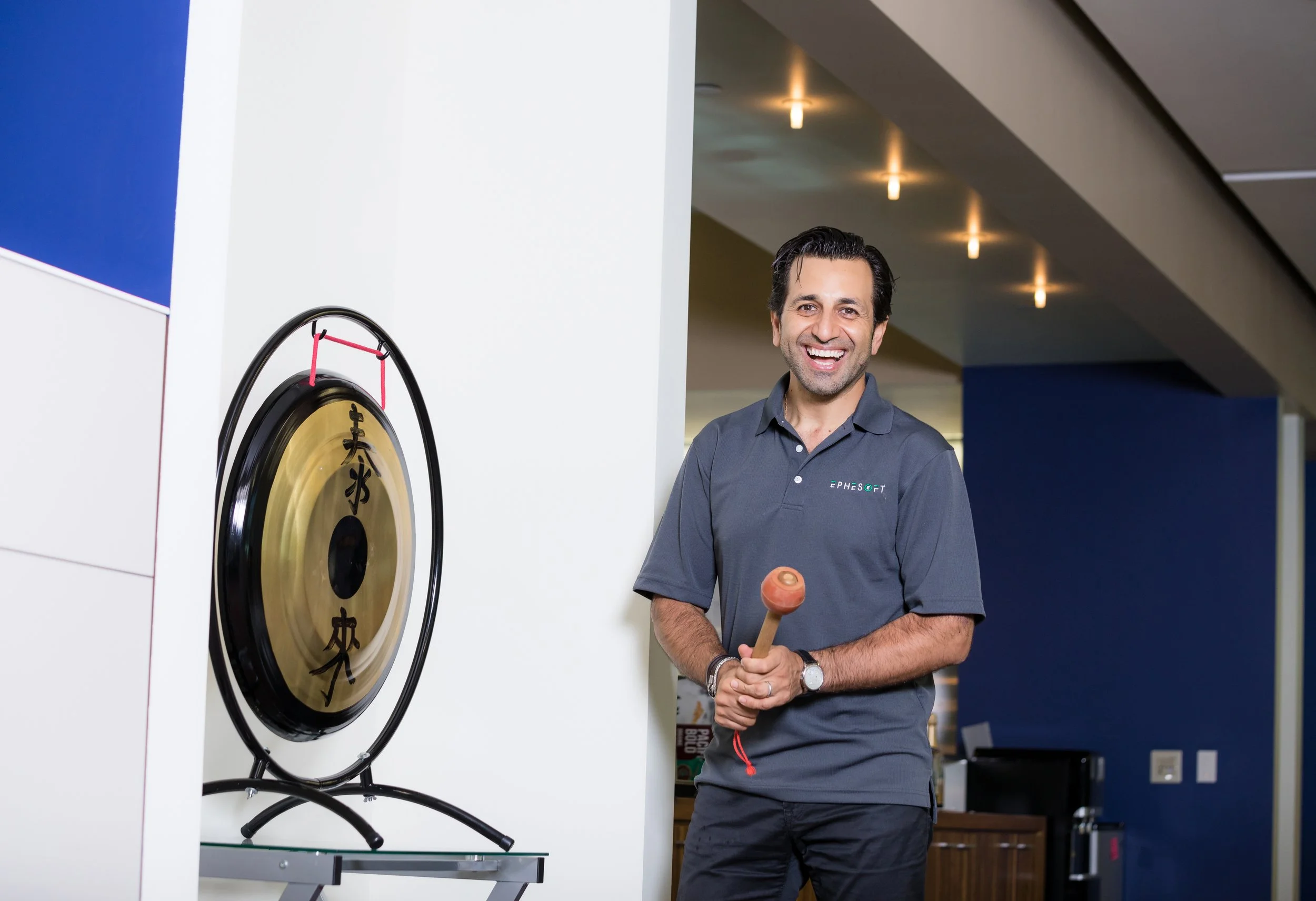A man smiling and holding a mallet, standing next to a large gong with Asian characters, in an indoor setting.