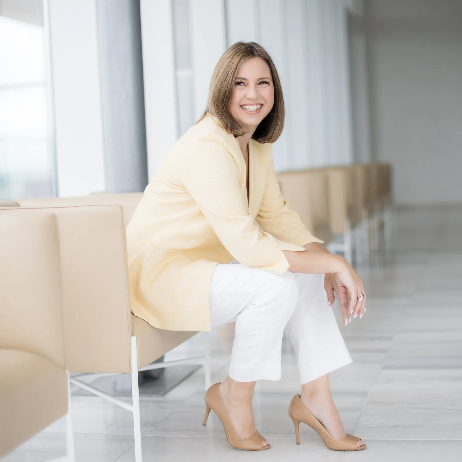 Personal branding photography of a woman in a pale yellow blazer seated in a bright Manhattan lobby