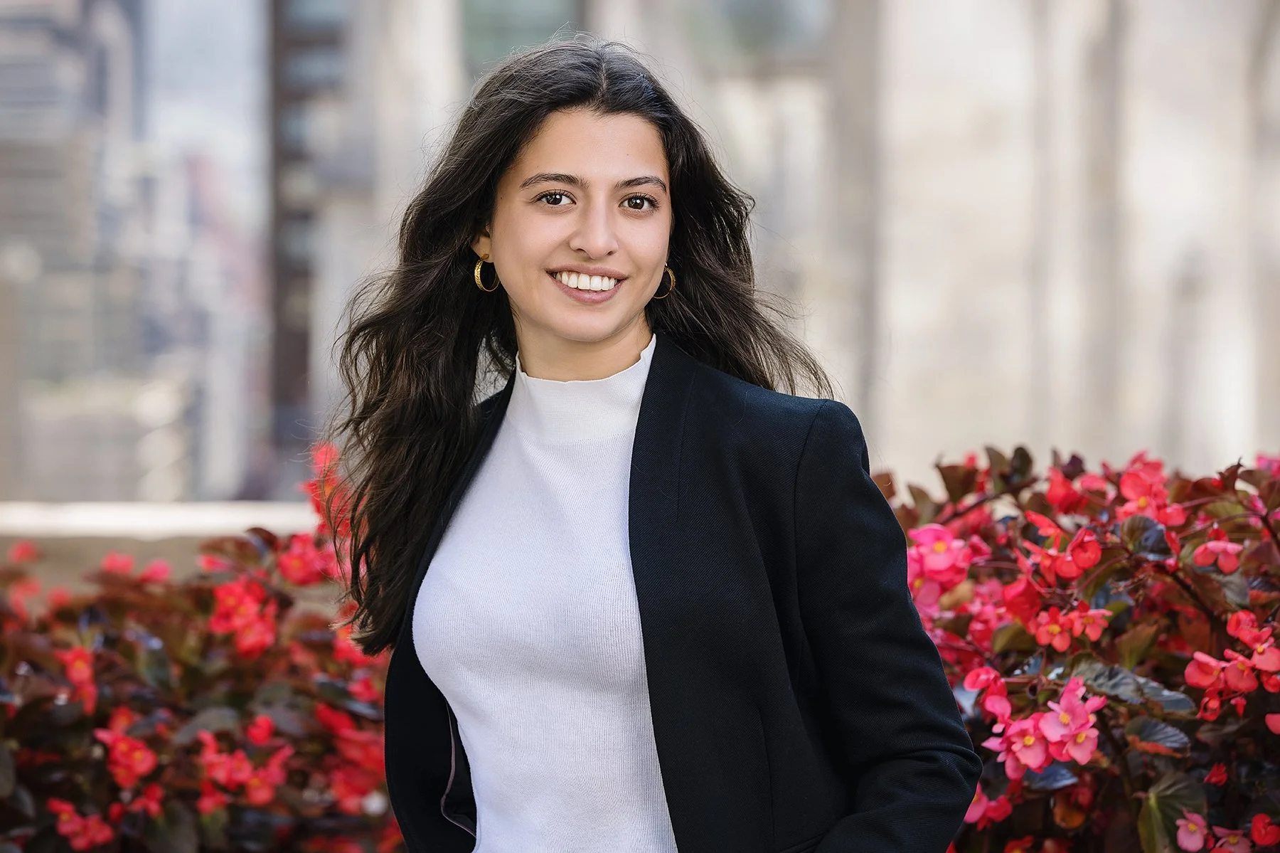 A young woman with long wavy dark hair, smiling, wearing a black blazer and white top, standing outdoors with pink flowers and blurred city buildings in the background.