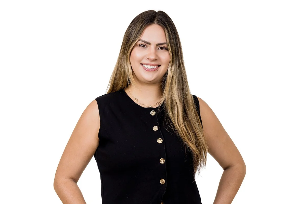 A woman with long light brown hair wearing a sleeveless black dress with gold buttons and a gold necklace, smiling at the camera against a white background.