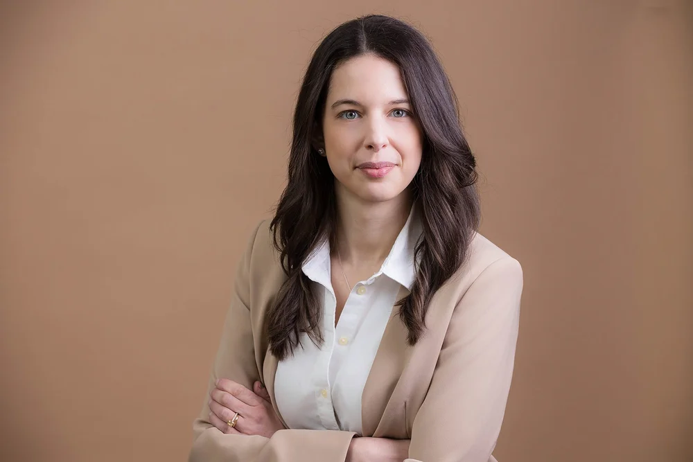 Consultant headshot of a woman in a beige blazer with arms crossed on a warm beige background