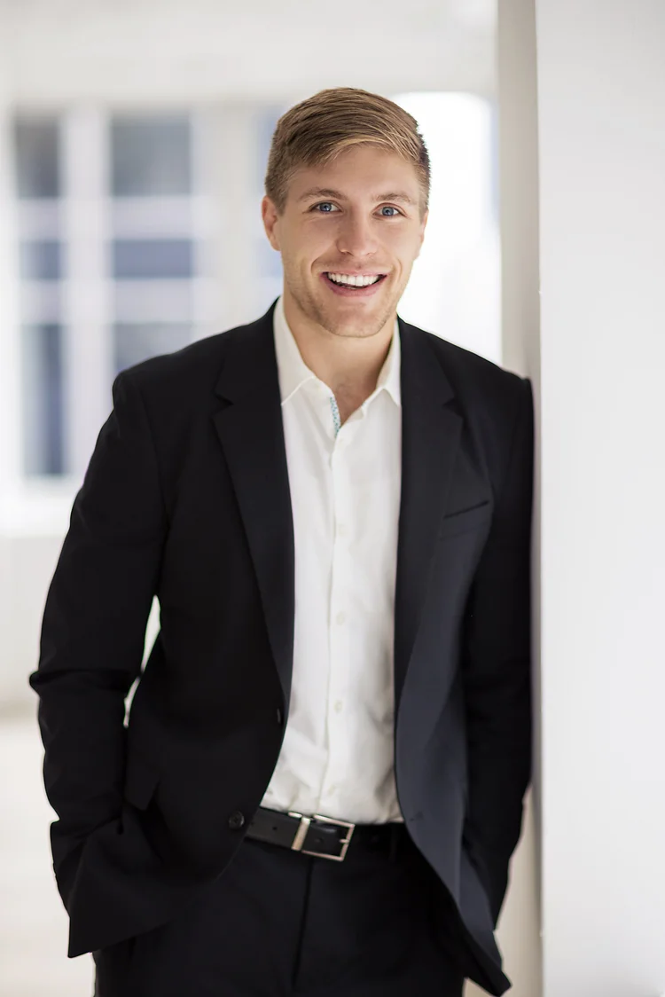 Office headshot of a smiling man in a black suit with hands in pockets by a bright window