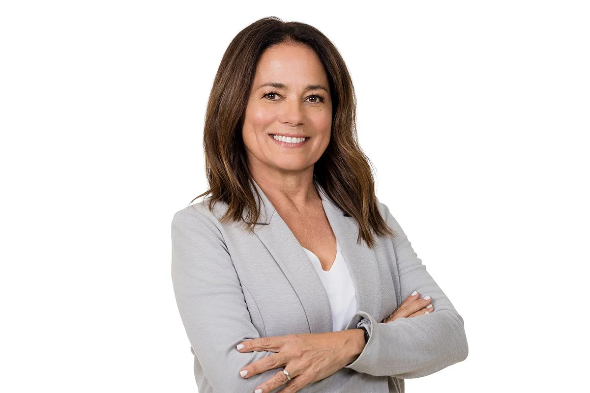 A woman with shoulder-length brown hair smiling and crossing her arms, wearing a light gray blazer over a white top, against a plain white background.