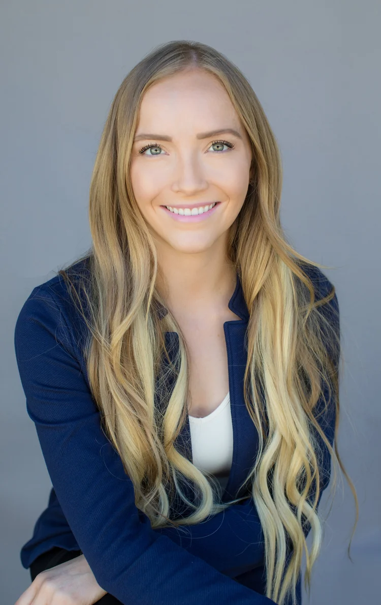 Studio headshot of a smiling woman with long blonde hair in a navy blazer on a grey background