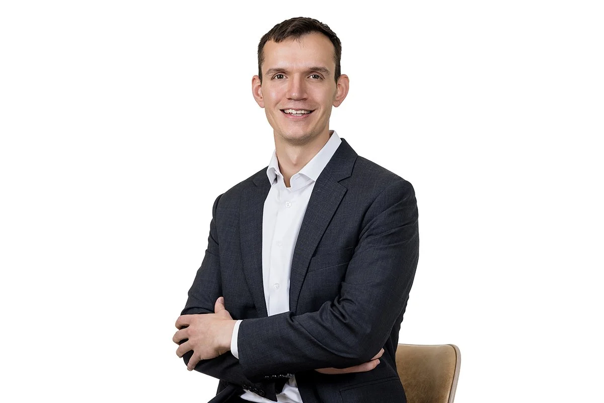 A young man with short dark hair, wearing a dark gray suit and white shirt, smiling and standing with arms crossed in front of a white background.