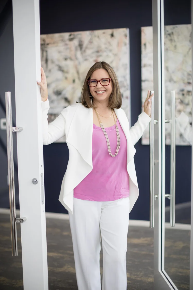 Personal branding portrait of a woman in a white blazer and pink top standing in an office doorway