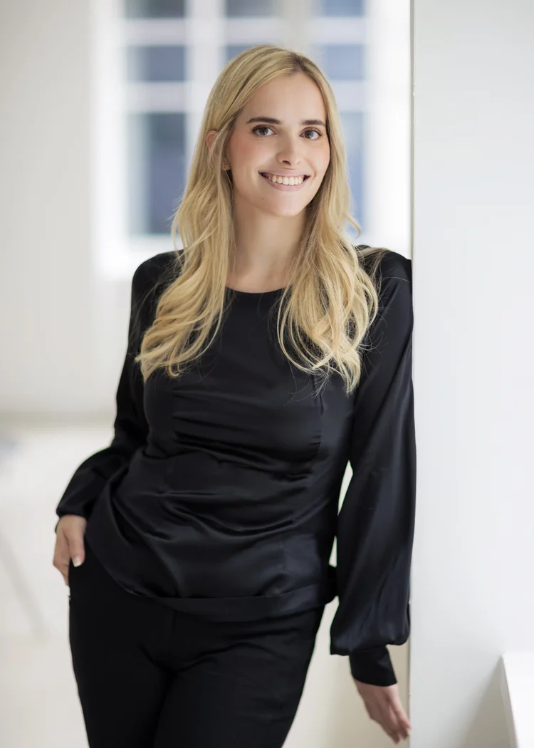 Executive headshot of a woman with long blonde hair in a black blouse leaning against a white wall