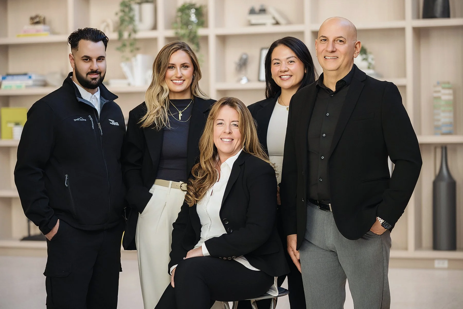 Six professionally dressed individuals posing together in an office setting with beige shelving in the background.