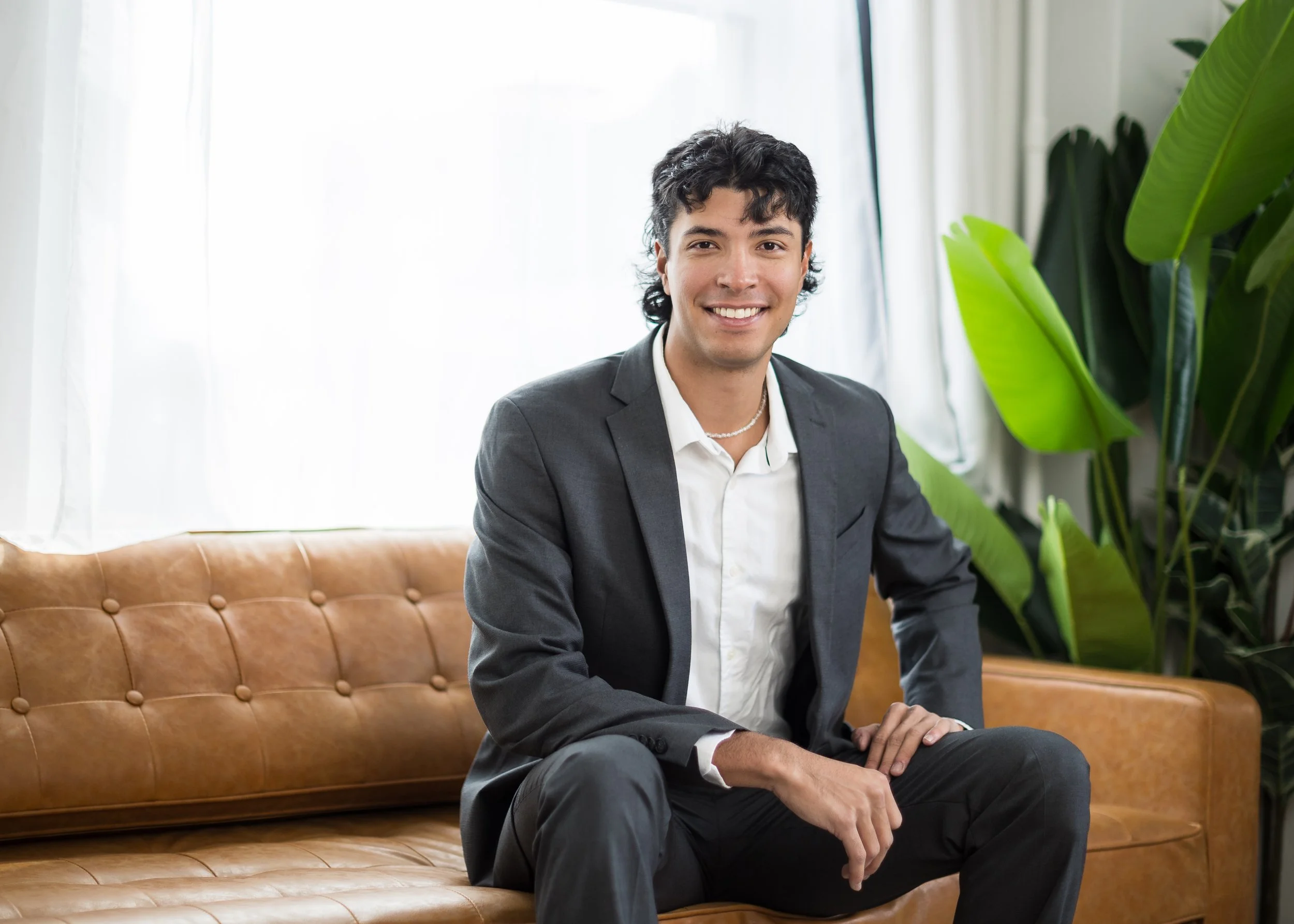 A smiling man in a gray suit and white shirt sitting on a brown leather couch in a bright room with large green plants.