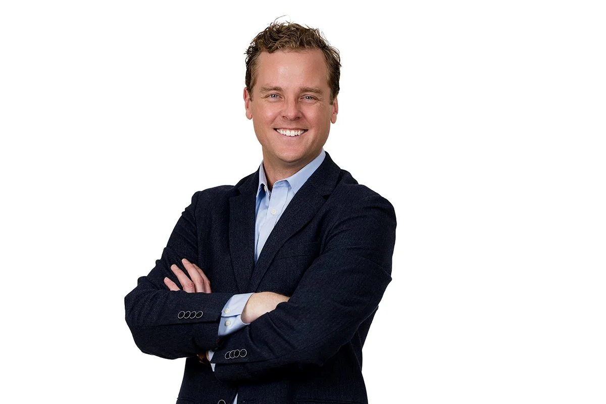 A smiling man with curly hair in a dark blazer and light blue shirt, arms crossed, against a plain white background.