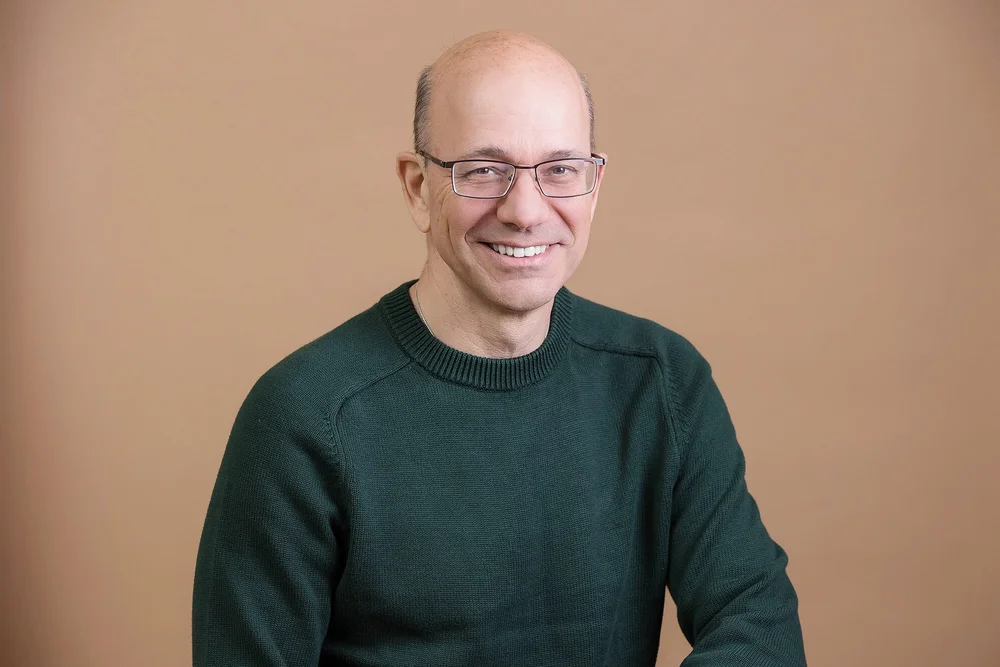Consultant headshot of a smiling man in glasses and a green sweater on a warm beige background