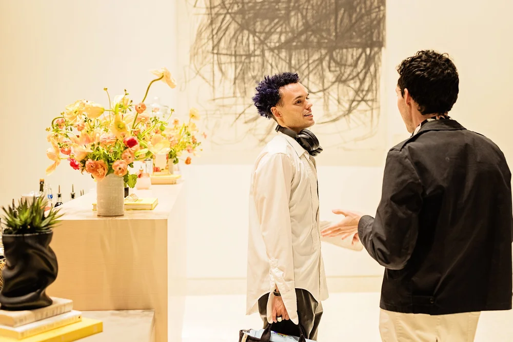 Two men in conversation in an art gallery with floral arrangements