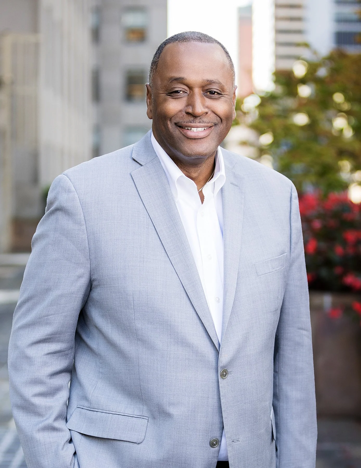 Professional man in light gray suit smiling outdoors, cityscape background with buildings and trees.