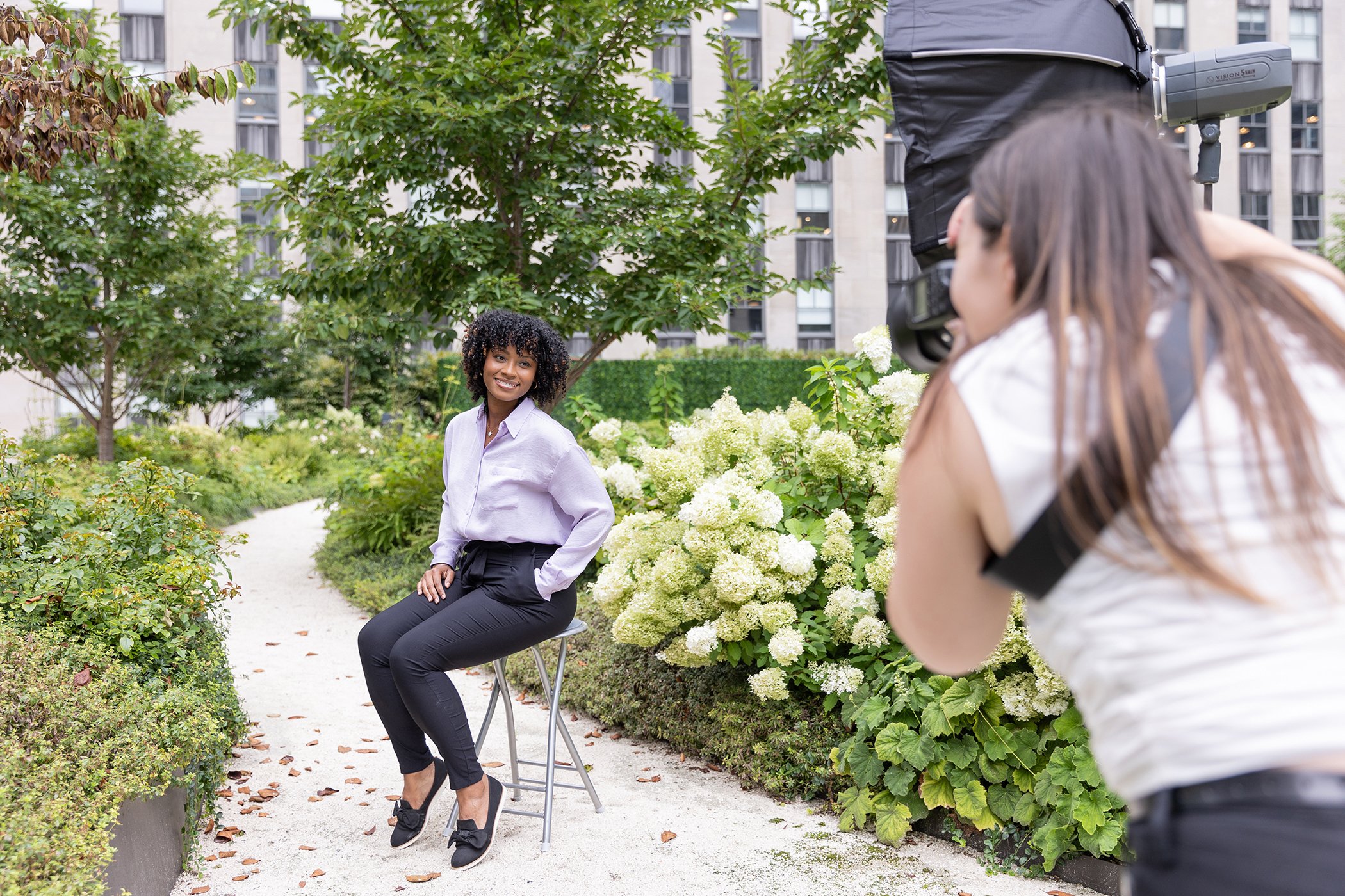 A woman is sitting on a stool in a garden, being photographed by a photographer with a professional camera. The woman is smiling and wearing a white blouse and black pants, surrounded by lush green plants and white flowers, with a cityscape in the background.