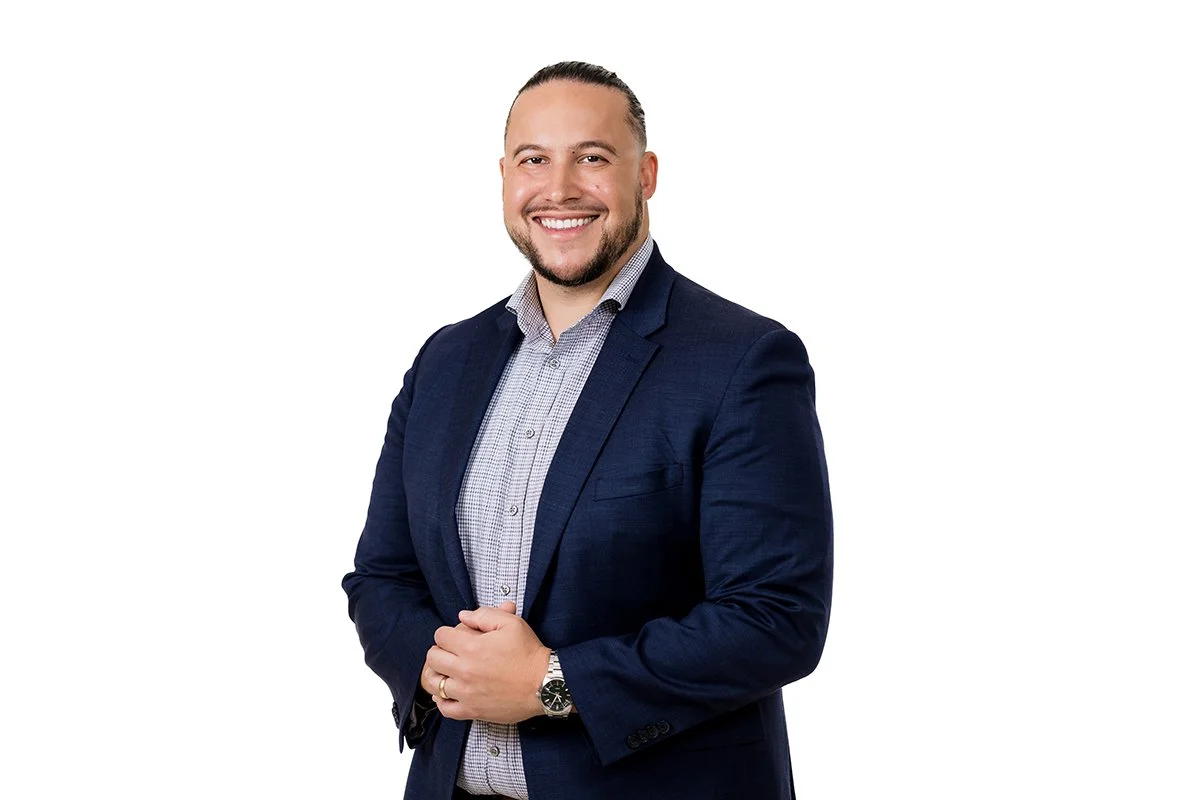 A man in a dark blue suit with a light checkered shirt, smiling and clasping his hands, against a white background.