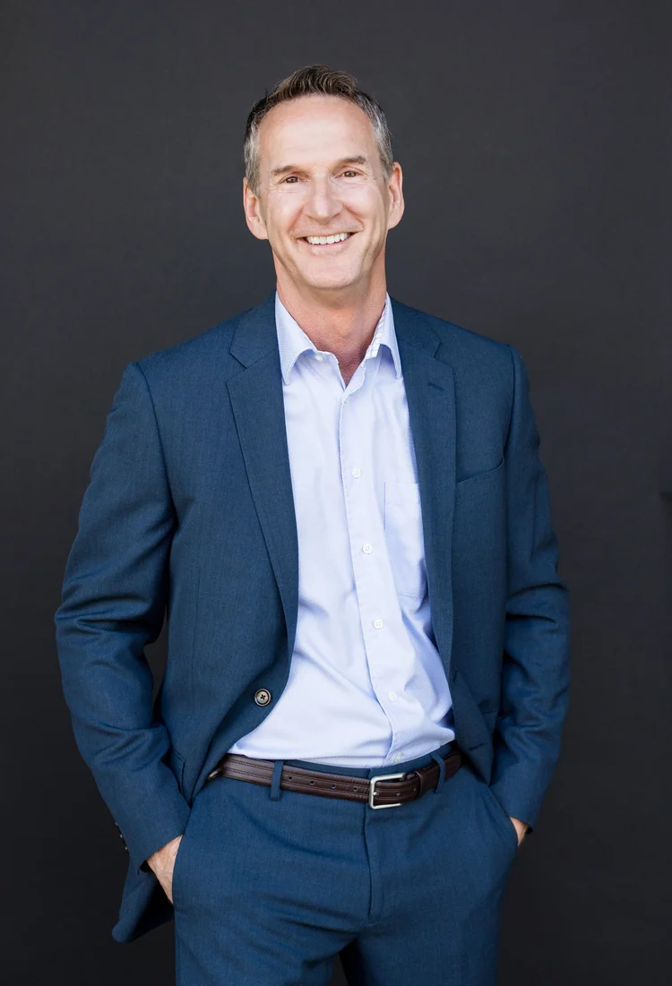 Executive coach headshot of a man in a blue suit with hands in pockets on a dark background