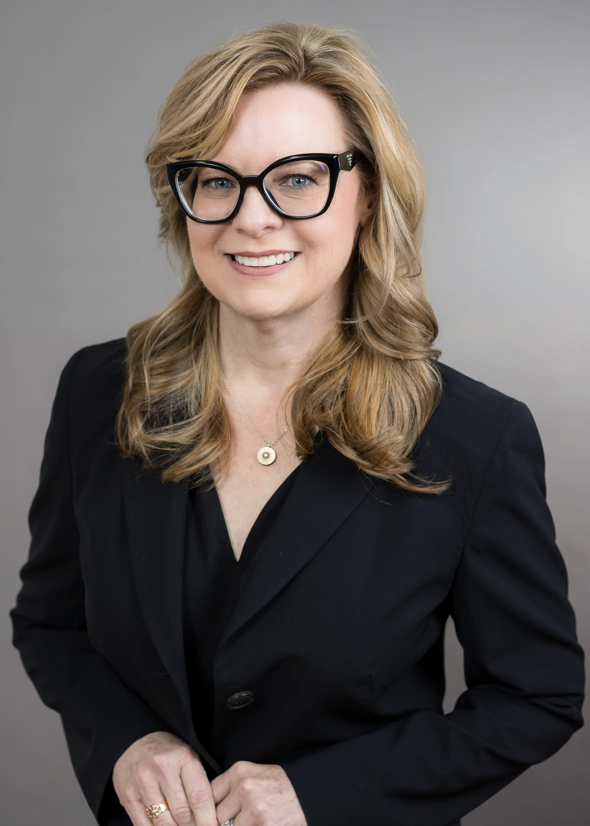 A woman with blonde hair wearing glasses, a black blazer, and a necklace, smiling against a gray background.