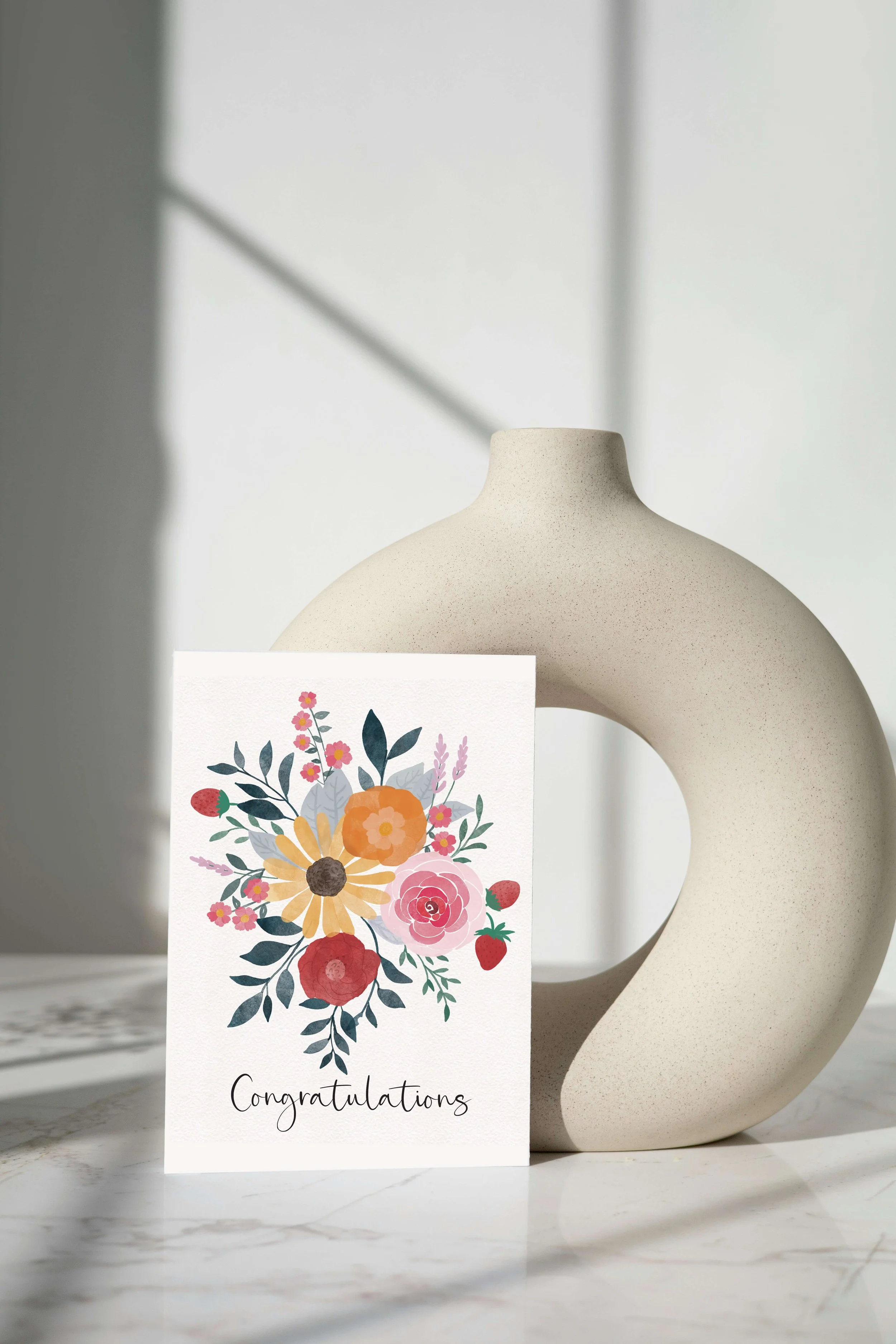 Greeting card that reads Congratulations with painted flowers at top. On a marble table with tan round vase