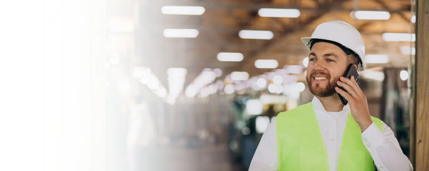 A man with a beard wearing a white hard hat and a yellow safety vest, talking on a cellphone inside a warehouse or construction site.