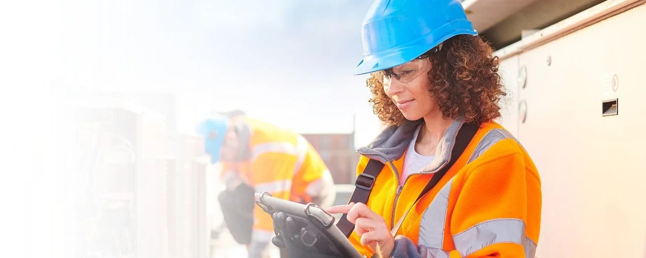 Woman construction worker wearing a blue safety helmet and orange safety vest, using a tablet on construction site with another worker in the background.