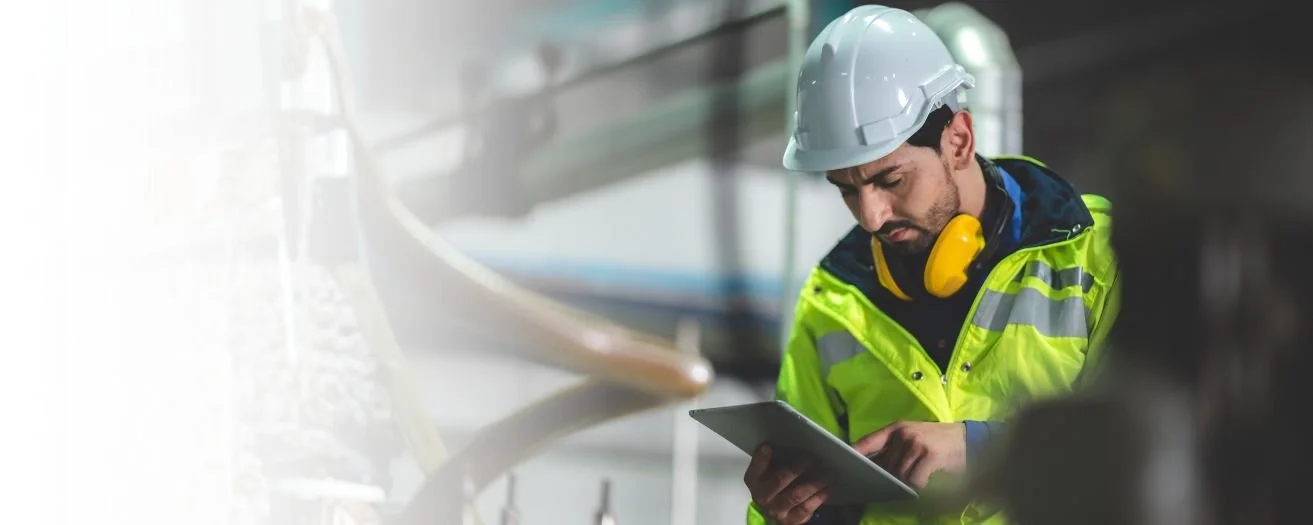 A man wearing a safety helmet and a high-visibility jacket, looking at a tablet in an industrial or construction setting.