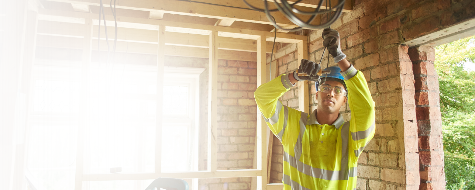 Construction worker installing electrical wiring in a building with exposed brick walls and wooden framing, wearing safety glasses, a blue helmet, and a high-visibility yellow shirt.