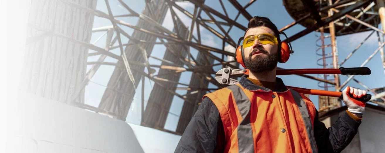 A construction worker with safety glasses, headphones, and a reflective vest holding a pipe wrench on top of a building under construction.