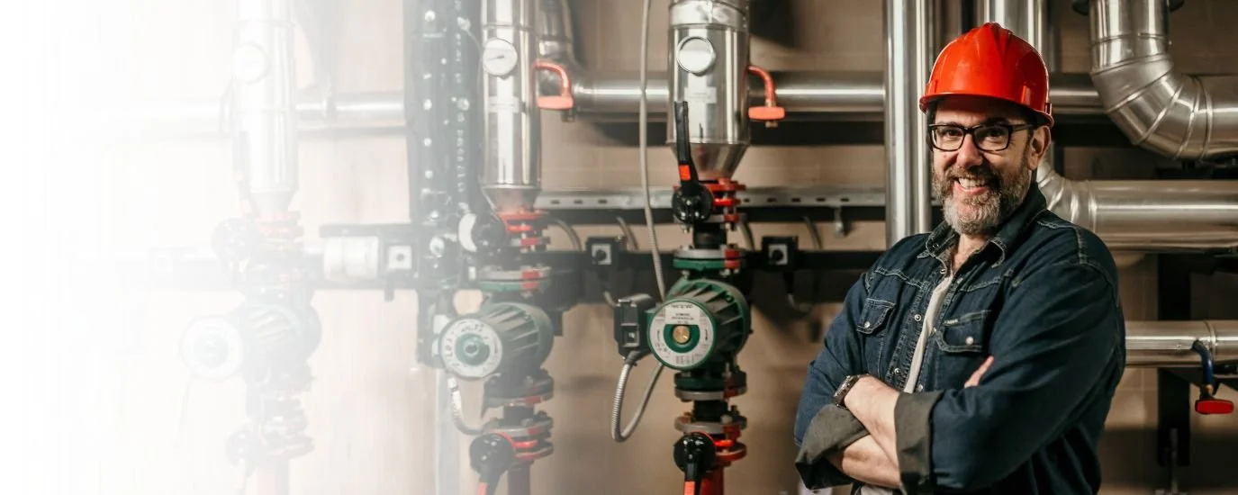 A man wearing safety glasses and a red safety helmet standing with arms crossed in front of industrial piping and valves in a mechanical room.