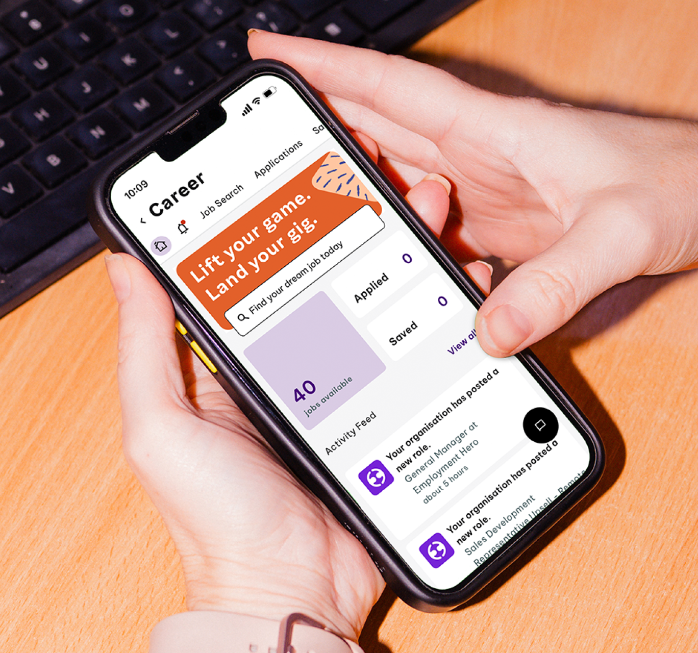 Person holding smartphone displaying a job search app on a wooden desk with a keyboard in the background.