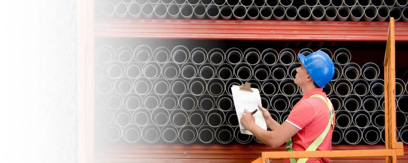 A construction worker wearing a blue safety helmet and a red shirt with yellow reflective straps is inspecting a shelf of black pipes in a warehouse, taking notes on a clipboard.