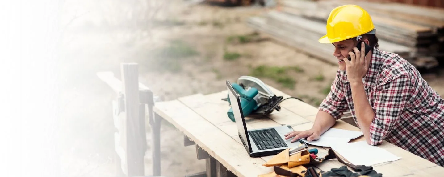 Construction worker in a yellow hard hat talking on a cell phone at a worksite, with a laptop and tools on a wooden table.