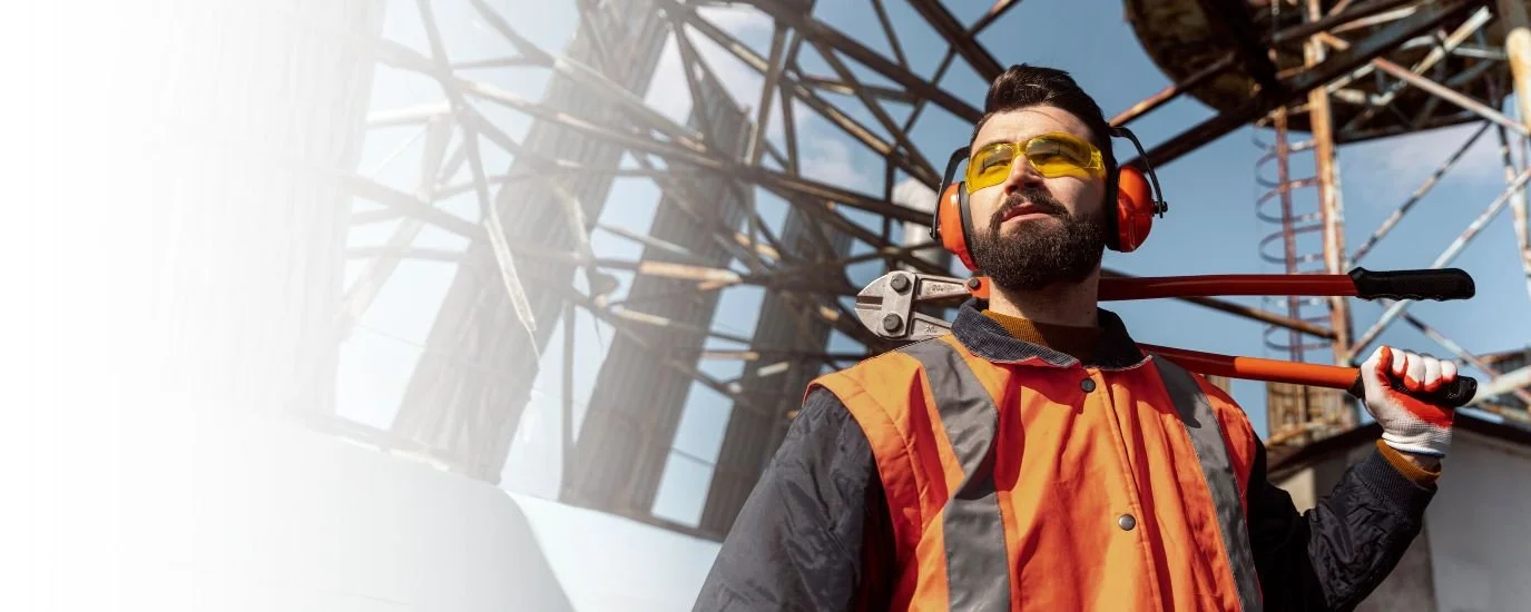 A construction worker with safety glasses and ear protection carrying a wrench on his shoulder at an industrial site with metal framework and a blue sky.