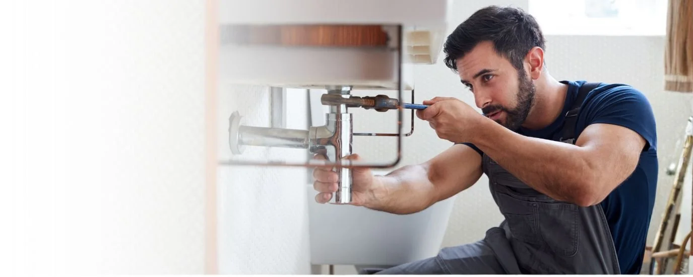 A man with dark hair and beard installing or fixing a plumbing pipe under a sink, using a screwdriver in a kitchen or bathroom setting.