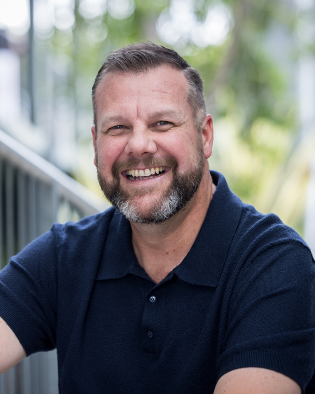 A smiling man with a beard, wearing a navy blue polo shirt, outdoors with blurred greenery in the background.