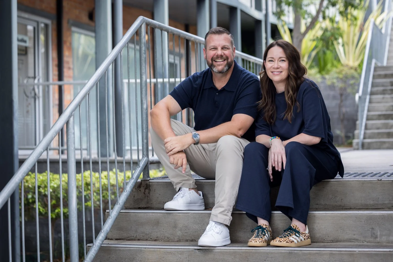 A man and woman sitting on outdoor stairs, smiling at the camera. The man has a beard and is wearing a navy polo shirt, beige pants, white sneakers, and a watch. The woman has long brown hair and is dressed in a navy top and black pants, with leopard print sneakers. There are plants and a building in the background.