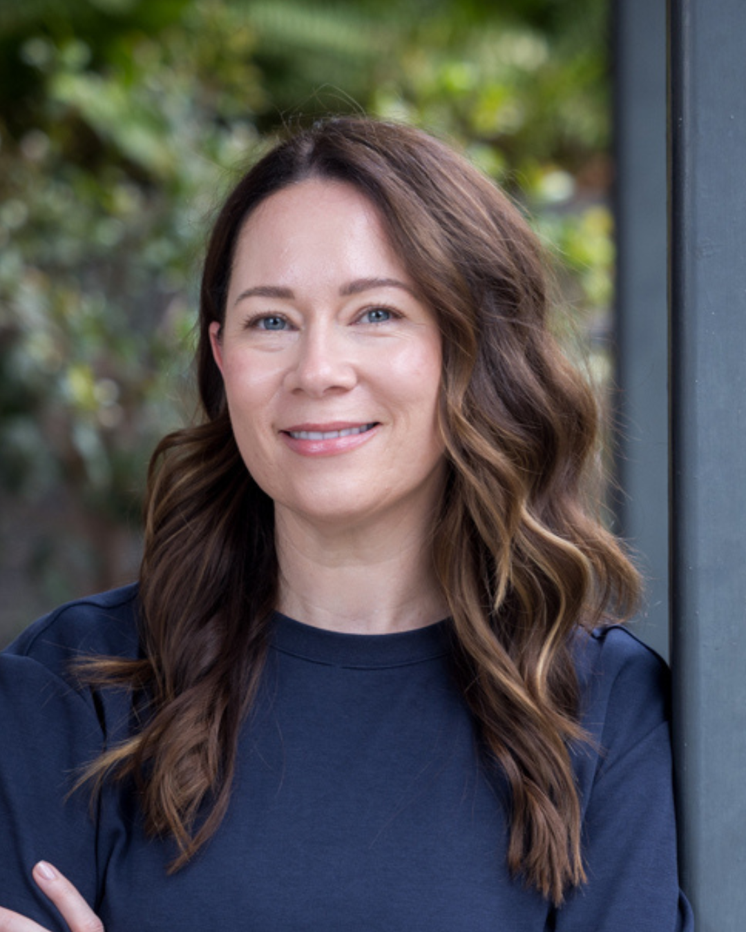 A woman with wavy brown hair and blue eyes smiling, standing outdoors next to a dark gray pillar with greenery in the background.