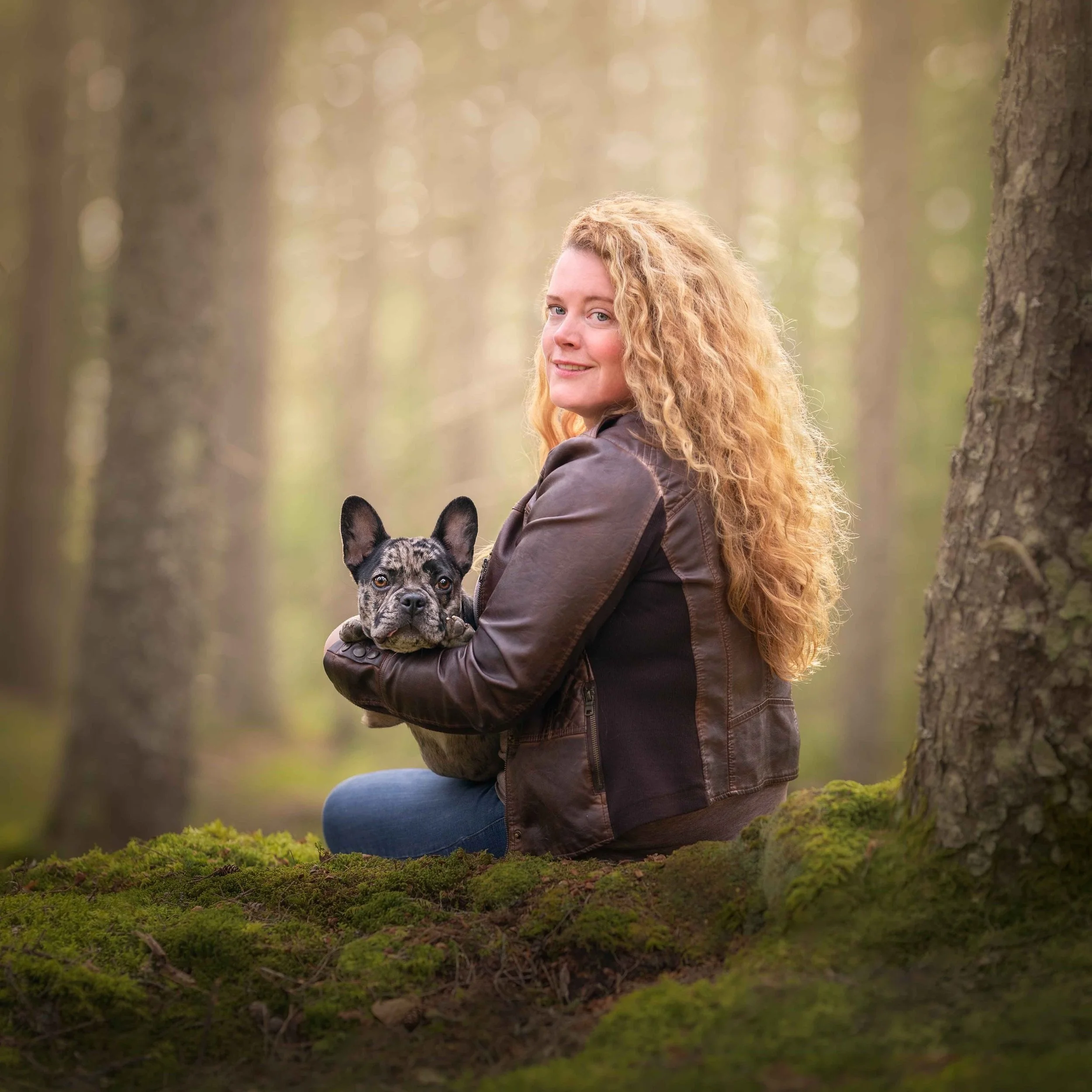 A woman with curly red hair sitting on mossy ground in a forest, holding a French bulldog puppy, both looking at the camera during professional dog photography session in freeport, maine.