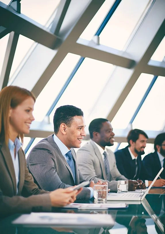 Business professionals sitting at a conference table in a modern, glass-walled room, engaged in a meeting or presentation.