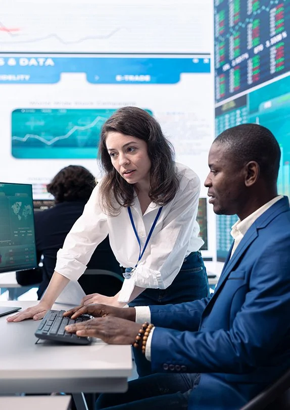 Two professionals working together at a computer in a high-tech office with financial data displayed on monitors and screens in the background.