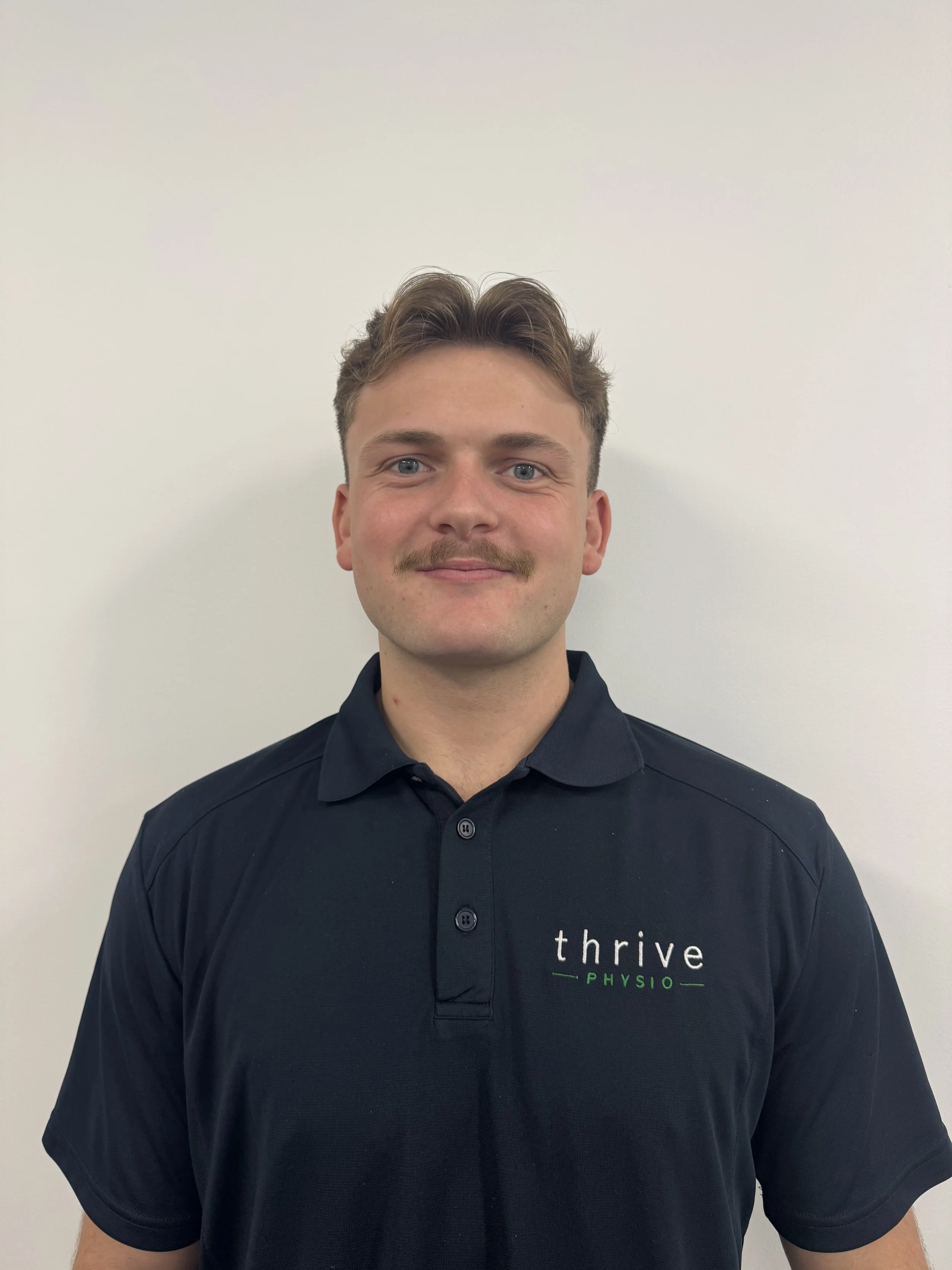 A young man with light skin, styled brown hair, blue eyes, and a mustache, wearing a black polo shirt with the words 'thrive PHYSIO' on it, standing against a plain white wall.